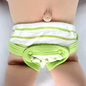 May include: A pair of white and lime green striped underwear featuring a solid lime green front panel with two matching snaps. The garment is worn on a doll torso, positioned below a small, round, beige belly button detail on the doll's skin.