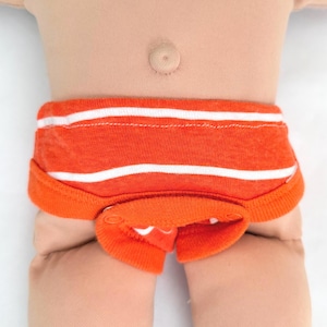 May include: A doll wearing orange underwear with horizontal white stripes. The garment features a ribbed waistband and a front flap secured with two orange snaps. The fabric has a textured appearance, and the design includes a solid orange trim.