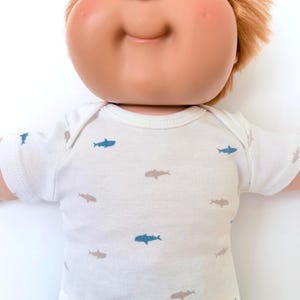 May include: A doll with a round face, bright blue eyes, and reddish-brown hair wearing a white short-sleeved bodysuit featuring a repeating pattern of small blue and light brown sharks. The doll has a smooth, light-toned complexion and a soft expression.