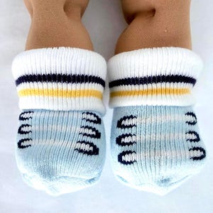 May include: A pair of light blue baby socks featuring a white ribbed cuff with horizontal navy blue and yellow stripes. The foot area displays a decorative pattern of three horizontal white lines on each side, accented by small navy blue stitched details.
