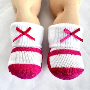 May include: A pair of baby socks with white ribbed cuffs and pink striped foot. Each sock has a small pink bow at the cuff. The socks are designed to fit a baby doll.