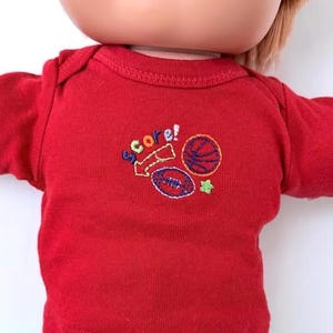 May include: A red short-sleeved onesie featuring an embroidered graphic on the chest. The design includes the word Score! in multicolored letters, a yellow arrow, a basketball, a football, and a small green star, worn by a doll.