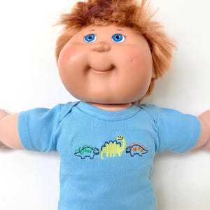 May include: A vintage doll with a vinyl face, blue eyes, and auburn hair. The doll wears a light blue short-sleeved shirt with embroidered dinosaurs in green, yellow, and orange. The doll has rosy cheeks and a slight smile.