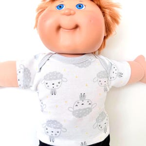 May include: A doll with red hair and blue eyes wearing a white onesie with a gray sheep pattern and yellow star accents. The doll has a smiling expression and is set against a white background.