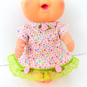 May include: A doll with a peach-toned complexion wearing a white dress featuring a small floral pattern in pink, blue, and yellow. The dress has a single pink rose at the neckline and two pink roses at the hem, which is finished with green lace.