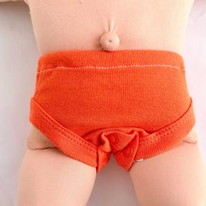 May include: A pair of bright orange ribbed knit underwear featuring a high waistband and a snap button closure at the crotch. The garment is worn on a light-toned doll body, highlighting the textured fabric and the visible white stitching detail.