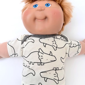May include: A doll with light skin, rosy cheeks, and short reddish-brown hair, featuring bright blue eyes. The doll wears a short-sleeved, cream-colored shirt with a repeating black line-art pattern of triceratops dinosaurs and a dark gray trim.