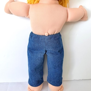 May include: A vintage doll with blonde yarn hair and a peach-colored body. The doll is wearing blue denim pants. The doll's arms are outstretched, and its feet are visible. The doll is a classic toy.