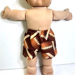 May include: A doll with light skin and light brown hair wearing brown shorts featuring a repeating pattern of tan and brown American footballs. The doll has outstretched arms, a rounded face, and a visible belly button on its soft-textured torso.