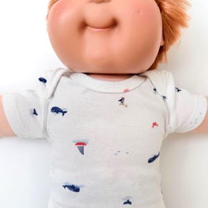May include: A doll with ginger hair and blue eyes wearing a white short-sleeved onesie. The onesie features a repeating pattern of small navy blue whales, red crabs, and birds perched on rocks, printed across the white fabric surface.