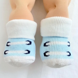 May include: A pair of baby socks with a white cuff and light blue foot. The foot of the sock has white stripes and dark blue decorative accents. The socks are on a white background.