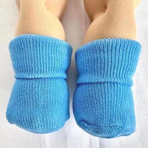 May include: A pair of bright blue knit booties worn on two feet. The booties feature a ribbed texture throughout the fabric and a folded cuff at the top, providing a snug fit around the ankles. The material appears to be a soft, woven yarn.