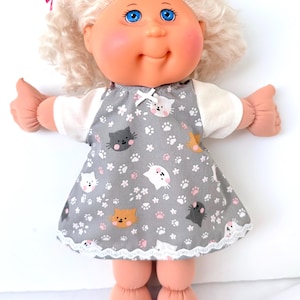 May include: A vintage doll with blue eyes, rosy cheeks, and blonde curly hair. The doll wears a gray dress with a white cat and paw print pattern, a white bow, and white lace trim. A pink ribbon is tied in the hair.