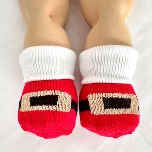May include: A pair of red knit socks with white ribbed cuffs, featuring a black horizontal band and a metallic gold rectangular buckle design on the front, worn on the lower legs of a doll with light skin tone and visible knee joints.
