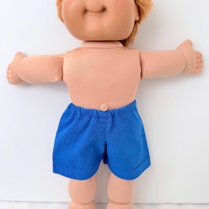 May include: A vintage doll with a vinyl head, soft body, and blue shorts. The doll has blue eyes, rosy cheeks, and light brown hair. The doll's arms are outstretched, and it is wearing blue shorts.
