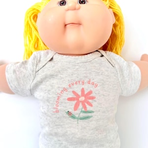 May include: A vintage doll with yellow yarn hair and a round face. The doll wears a gray onesie with a pink flower and the words "blooming every day". The doll's eyes are brown and it has a slight smile.