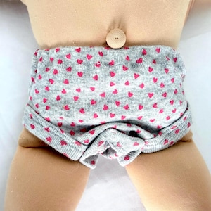 May include: A pair of heather gray underwear featuring a repeating pattern of small pink hearts. The garment is made of a ribbed knit fabric and includes a single white snap button closure at the center front, worn on a light-toned doll body.