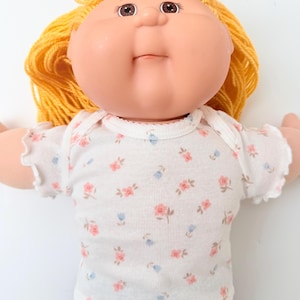 May include: A doll with a round, peach-toned face, brown eyes, and bright yellow yarn hair. The doll wears a white short-sleeved shirt featuring a small, repeating floral pattern in pink and light blue, with ruffled edges on the sleeves.