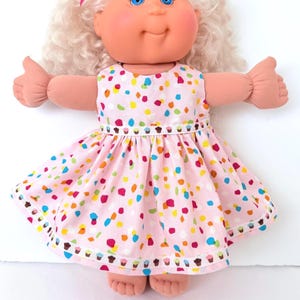 May include: A doll with light skin, blue eyes, and curly blonde hair wearing a light pink dress. The dress features a colorful confetti pattern and a white ribbon trim decorated with small cupcake graphics at the waist and the hemline.