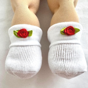 May include: A pair of white ribbed socks with a red rose and green leaf embellishment. The socks are worn on beige fabric limbs, likely part of a doll or craft project. The socks are made of a soft, knitted material.