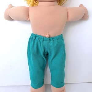 May include: A doll with light skin, a prominent belly button, and yellow yarn hair. The doll is wearing solid teal pants with an elastic waistband. The doll has outstretched arms and visible toes on both feet, presented against a plain background.