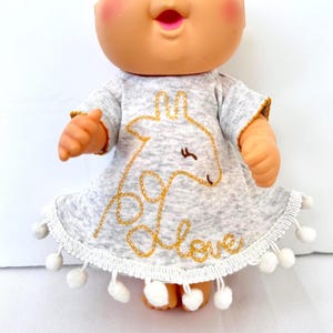 May include: A doll with a round head, blue eyes, and pink cheeks wearing a heather gray short-sleeved dress. The dress features a gold embroidered giraffe outline and the word love in cursive, with white pom-pom trim along the bottom hem.
