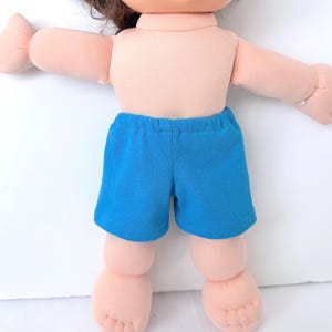 May include: A doll with light skin and brown hair wearing bright blue shorts. The doll has a soft fabric body with rounded limbs, visible fingers, and toes. The shorts feature an elastic waistband and a simple hemmed design on the legs.