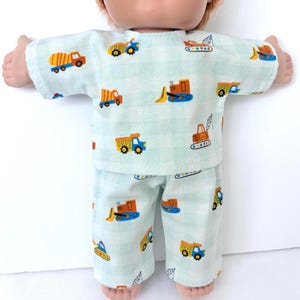 May include: A doll wearing a light blue pajama set with a repeating pattern of colorful construction vehicles. The set includes a short-sleeved top and matching pants. The vehicles are orange, yellow, blue, and red.