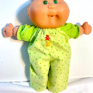 May include: A doll with a round peach-colored head, green eyes, and a small mouth. The doll wears a light green long-sleeved jumpsuit featuring a pattern of small white and dark green dots, accented by two small bear-shaped buttons in red and yellow.