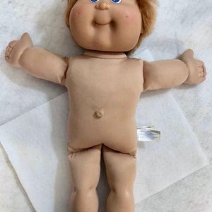 May include: A doll with a soft fabric body, light skin tone, and bright blue eyes. The doll features short, messy reddish-brown hair, a small belly button, and outstretched arms. A white fabric tag is attached to the side of the torso.