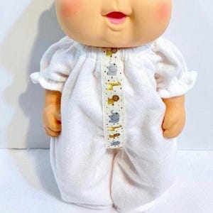 May include: A doll wearing a white onesie with a decorative ribbon down the front. The ribbon features animal illustrations, including elephants and lions. The doll has rosy cheeks and a smiling expression.