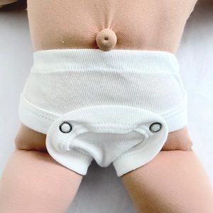 May include: A close-up of a doll wearing a white onesie. The onesie has a snap closure and is made of a soft, textured fabric. The doll's skin is a light peach color.