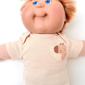 May include: A doll with a round face, bright blue eyes, and messy reddish-brown hair. The doll wears a short-sleeved, beige and white horizontally striped shirt featuring a small chest pocket with an embroidered brown and tan giraffe head.