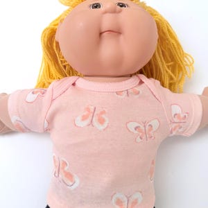 May include: A doll with a round face, tan skin, and yellow yarn hair. The doll wears a short-sleeved, light pink shirt featuring a repeating pattern of white and orange butterflies. The doll has dark brown eyes and a small, closed mouth.