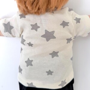 May include: A short-sleeved t-shirt in an off-white color featuring a repeating pattern of gray stars of varying sizes across the front and sleeves. The garment has a crew neckline and is worn by a figure with reddish-brown hair visible at the top.