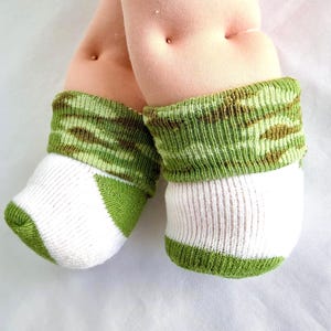 May include: A pair of baby socks featuring a white foot section with green accents and a folded cuff in a green and brown camouflage pattern. The socks are worn on soft, light-toned doll feet that have small indentations at the ankle area.