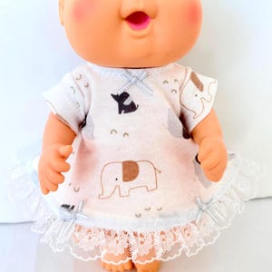 May include: A Cabbage Patch Kid doll wearing a light pink and white dress with elephant and crescent moon patterns. The dress has white lace trim and small silver bows. The doll has blue eyes and rosy cheeks.