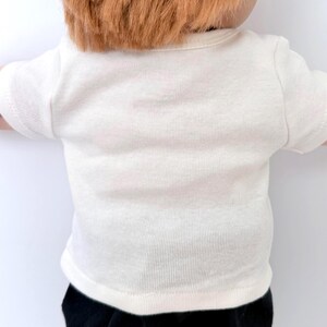 May include: A short-sleeved, off-white ribbed t-shirt with a crew neckline. The garment is worn by a doll with reddish-brown hair. The fabric has a visible vertical texture, and the shirt features a simple, plain design with hemmed sleeves.