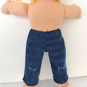 May include: A doll with light skin and yellow yarn hair wearing dark blue denim pants. The pants feature a distressed look with frayed hems and small rips at the knees. The doll has a visible belly button and is shown from the waist down to the feet.