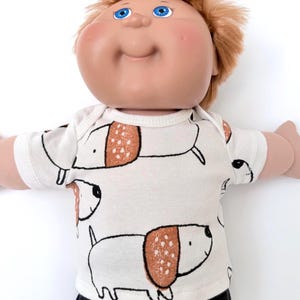 May include: A doll with light skin, blue eyes, and short reddish-brown hair wearing a white short-sleeved shirt featuring a repeating pattern of stylized white and brown dogs with dotted ears, paired with black pants.