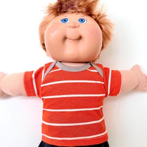 May include: A doll with a round face, rosy cheeks, and bright blue eyes, featuring messy light brown hair. The doll is wearing a short-sleeved, orange t-shirt with thin white horizontal stripes and a grey ribbed neckline, paired with black pants.