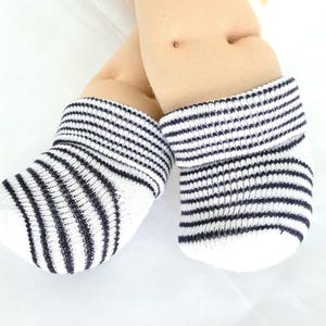 May include: A pair of baby booties featuring a white base with thin horizontal navy blue stripes. The booties have a folded cuff at the top and are worn on two soft, light-toned doll feet. The knit fabric shows a consistent striped pattern.