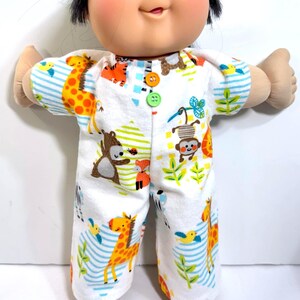 May include: A Cabbage Patch Kid doll wearing white pajamas with a colorful jungle animal print. The pajamas feature a pattern of giraffes, monkeys, and other animals. The doll has black hair and rosy cheeks.