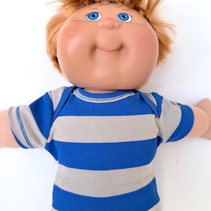 May include: A doll with a round, tan-colored face, bright blue eyes, and messy reddish-brown hair. The doll is wearing a short-sleeved shirt featuring horizontal stripes in alternating shades of royal blue and light gray, paired with black pants.
