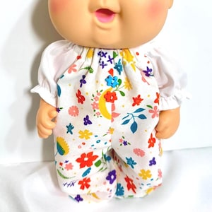 May include: A doll with a fair complexion and rosy cheeks wearing a white top and floral print jumpsuit. The jumpsuit features a white background with colorful flowers, a crescent moon, and blue, red, yellow, and purple accents. The doll's top has short sleeves.