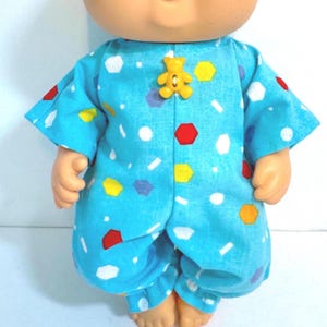 May include: A doll wearing a light blue romper featuring a pattern of small white, red, yellow, and purple geometric shapes. The garment is fastened at the center chest with a small, yellow, bear-shaped button. The doll has a round face.
