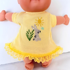 May include: A doll with a peach-toned complexion wearing a short-sleeved, ribbed yellow dress. The dress features an embroidered design of a gray rabbit, green grass, a yellow sun, and a small blue butterfly, finished with a yellow lace trim.