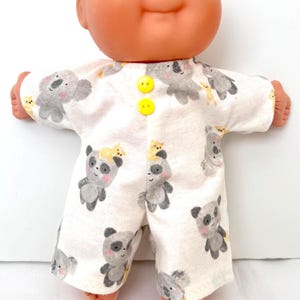 May include: A doll with a round peach-toned head, blue eyes, and blonde hair, wearing a white short-sleeved romper featuring a repeating pattern of gray koalas and pandas with small yellow accents, secured by two bright yellow buttons at the neck.