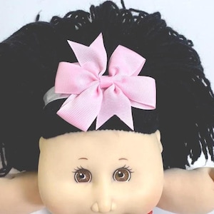 May include: A Cabbage Patch Kids doll with black yarn hair and a light pink bow. The doll has brown eyes and a light skin tone. The doll is wearing a bib with text.
