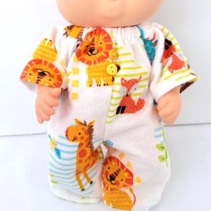 May include: A doll wearing a white onesie with a colorful animal print. The print features lions, giraffes, and foxes. The onesie has yellow stripes and buttons. The doll's face is visible.